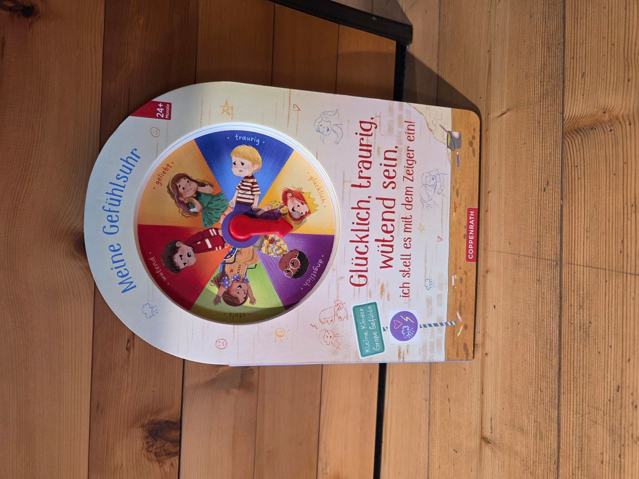 A colorful board book titled 'Meine Gefühlesuhr' with a spinning wheel showing various emotions. The wheel is labeled with different emotions like happy, sad, angry, and surprised. The book is placed on a wooden surface.