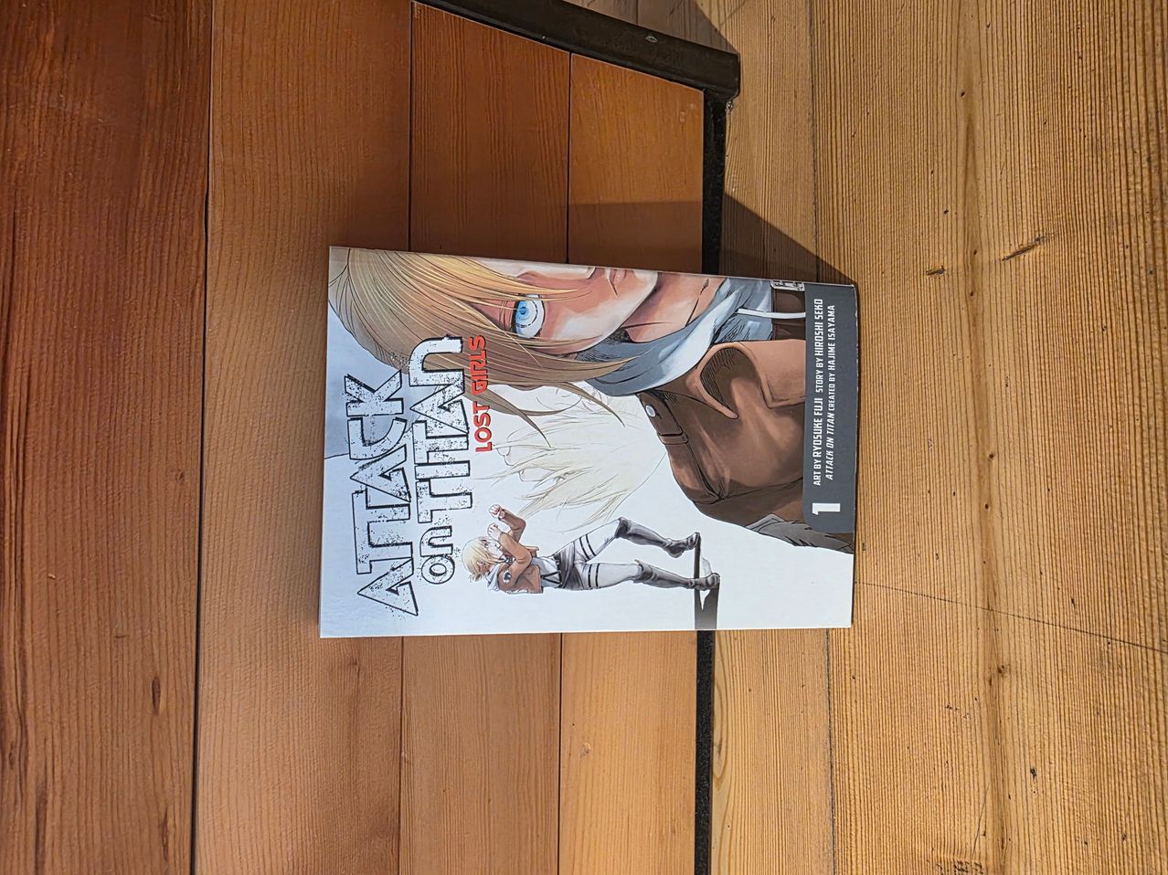 A manga book titled Attack on Titan is placed on a wooden surface. The cover features a character in a fighting stance with another character in the background.