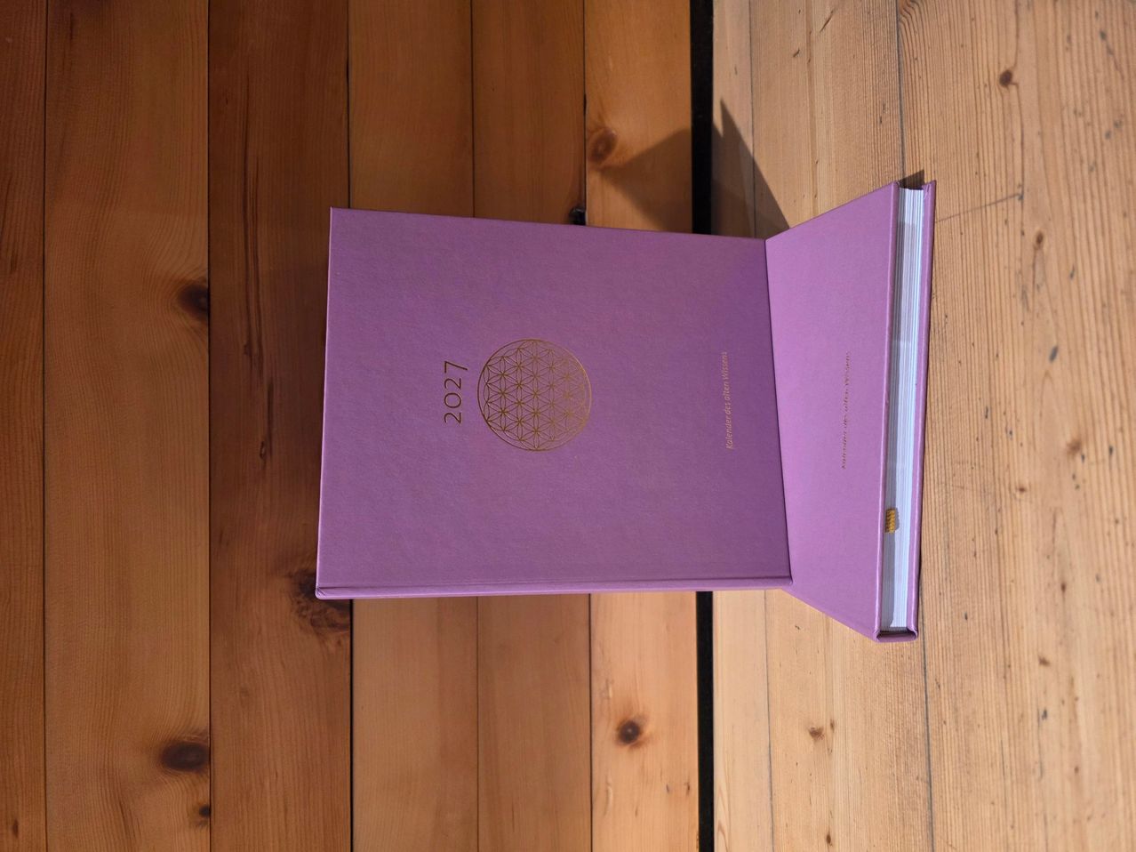 A purple book with the year 2027 on the cover, opened on a wooden surface. The cover features a golden flower of life design.