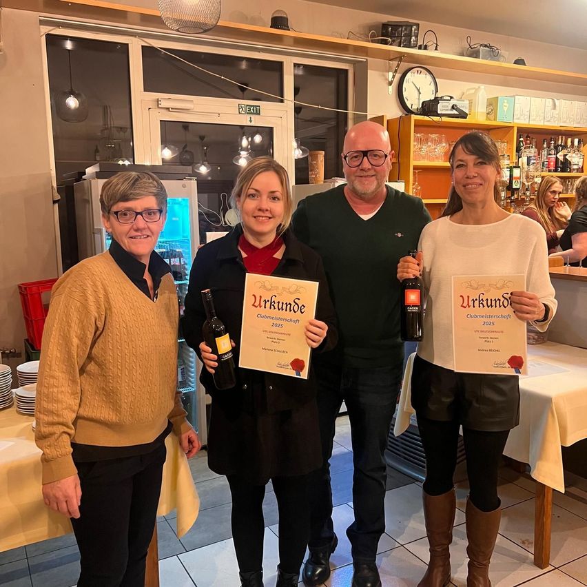 Drei Personen stehen in einem Restaurant mit Urkunden und Weinflaschen. Ein Mann und zwei Frauen. Dahinter steht ein Tisch mit Tellern.
