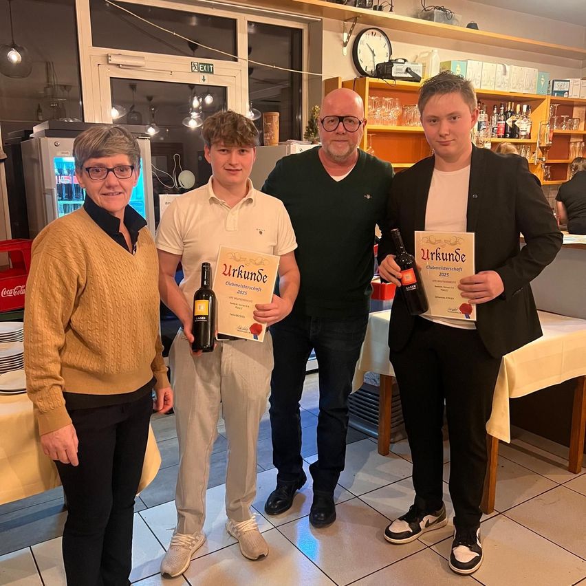 Vier Personen stehen in einem Raum und posieren für ein Foto. Drei von ihnen halten Urkunden und Weinflaschen in der Hand. Ein Kühlschrank steht hinter ihnen, und ein Regal mit Flaschen und anderen Gegenständen befindet sich hinter dem Mann in der Mitte. Eine Uhr ist über dem Regal an der Wand montiert.