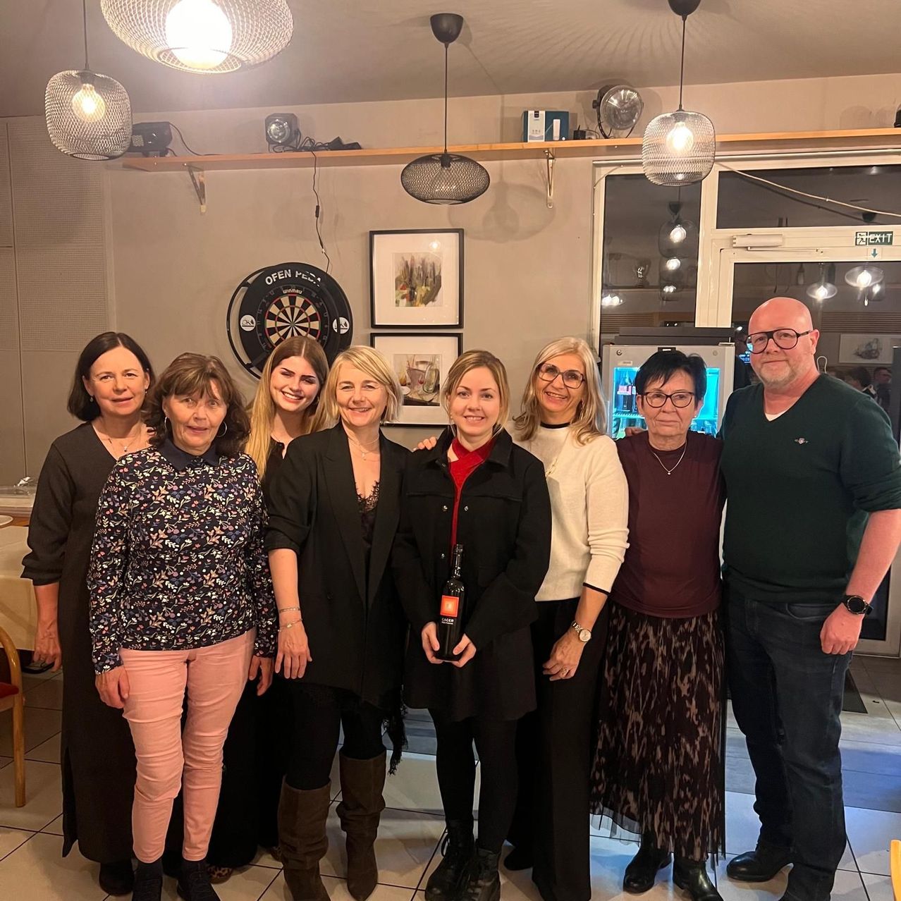 Sieben Menschen stehen zusammen in einem Raum. Drei Frauen halten Weinflaschen. Ein Dartboard und Bilderrahmen sind an der Wand.