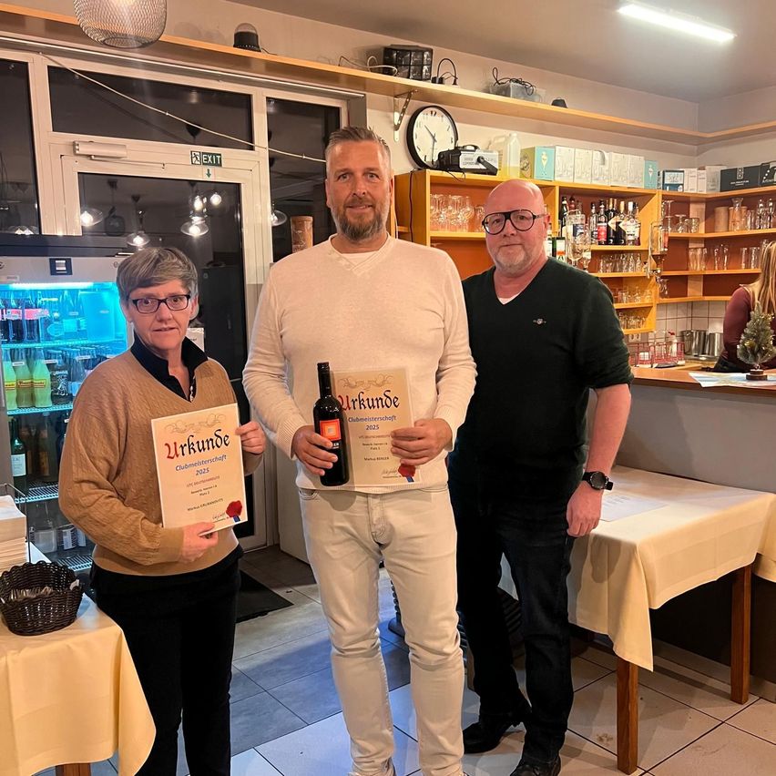 Drei Personen stehen in einem Raum und halten Urkunden und eine Flasche. Der Mann in der Mitte hält eine Weinflasche, und sie scheinen alle zu feiern. Hinter ihnen sind Regale mit Gläsern und Flaschen zu sehen.