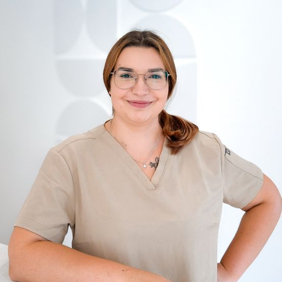 Eine Frau in einem beigen Scrub-Oberteil, das eine Brille und eine Halskette trägt, posiert lächelnd für ein Foto.