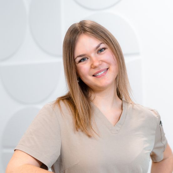 Eine Frau mit blonden Haaren lächelt und trägt ein hellbraunes V-Ausschnitt-Shirt, mit einem weißen geometrischen Hintergrund hinter ihr.