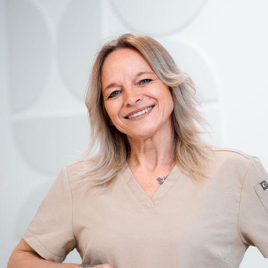 Eine lächelnde Frau mittleren Alters mit blonden Haaren in einem beigen Scrub-Anzug, vor einem weißen Hintergrund stehend.