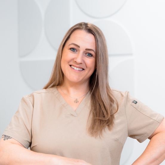 Eine Frau mit langen blonden Haaren, die ein beigefarbenes Uniformhemd trägt, lächelt herzlich mit verschränkten Armen vor einem weißen Hintergrund.