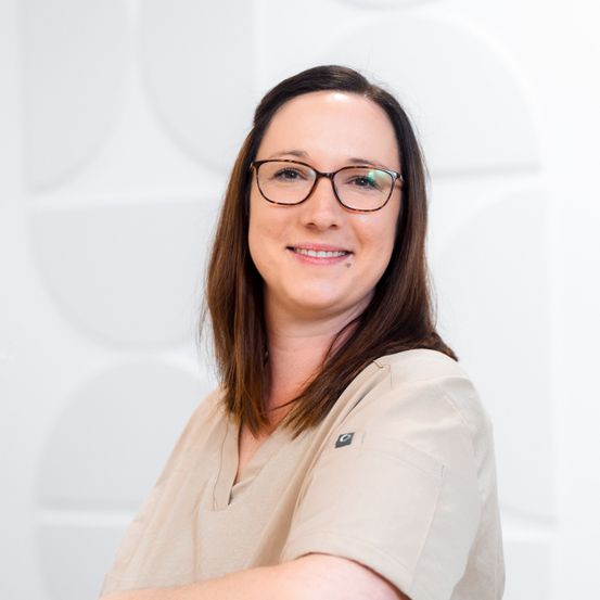 Eine Frau mit braunen Haaren und Brille lächelt für ein Porträt in einem beigen Hemd vor weißem Hintergrund.