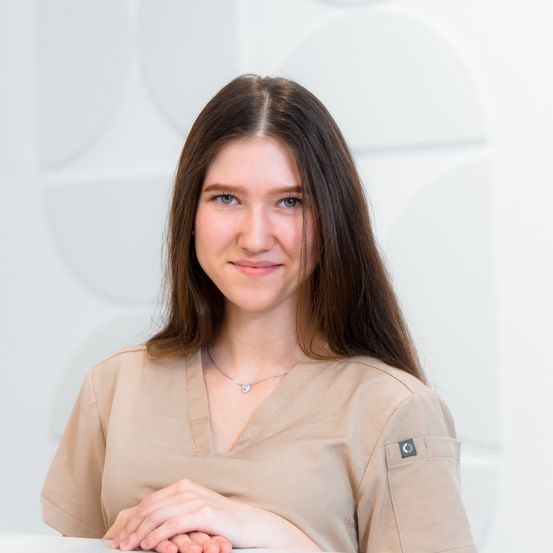 Eine Frau mit braunen Haaren in einem beigen Uniform lächelt und posiert für ein Foto vor einem weißen Hintergrund.
