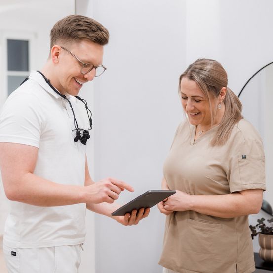 Ein Mann und eine Frau in medizinischen Uniformen lächeln und schauen auf ein Tablet in einem Büro.