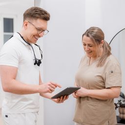 Ein Mann und eine Frau in medizinischen Uniformen lächeln und schauen auf ein Tablet in einem Büro.