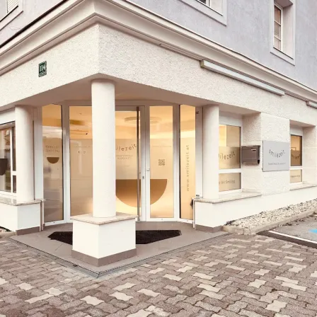 Der Eingang eines modernen Gebäudes, 'ilezeit', mit Glastüren und Fenstern, einem gepflasterten Gehweg und einem kleinen Schild an der Wand.