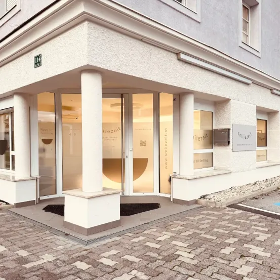 Der Eingang eines modernen Gebäudes, 'ilezeit', mit Glastüren und Fenstern, einem gepflasterten Gehweg und einem kleinen Schild an der Wand.