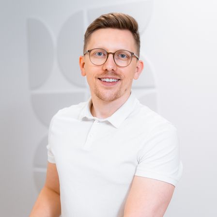 Ein Mann mit einem weißen Poloshirt und einer Brille lächelt und posiert für ein Foto. Er steht vor einer weißen Wand mit geometrischen Mustern.