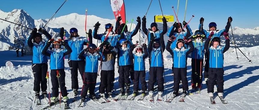 Eine Gruppe von Skifahrern in blauen Jacken posiert für ein Foto auf einem verschneiten Berg, mit Skistöcken und Helmen, unter einem klaren Himmel mit Bergen im Hintergrund.