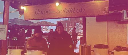 A snowy food stand named Union-SchiikLub with two people inside. There are pots and bottles on the table and people standing around.