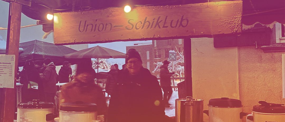Ein verschneiter Essensstand namens Union-SchiikLub mit zwei Personen im Inneren. Es gibt Töpfe und Flaschen auf dem Tisch und Leute, die herumstehen.