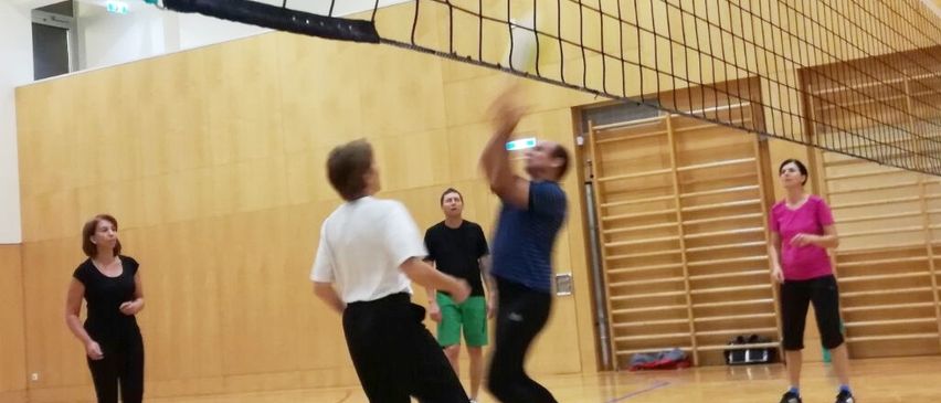 Eine Gruppe von Menschen spielt Volleyball in einer Halle. Ein Spieler springt, um den Ball zu schlagen, während ein anderer Spieler sich darauf vorbereitet, zu blocken. Zuschauer beobachten von den Seitenlinien.