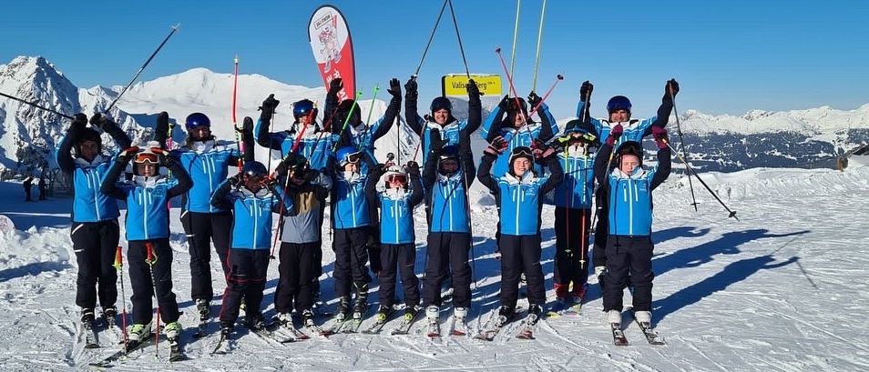 A group of people in blue ski gear pose on a snowy mountain, holding ski poles, with mountains and a sign in the background.
