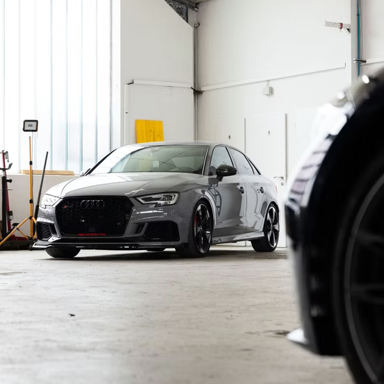 Ein grauer Audi RS 5 steht in einer Garage mit weißen Wänden. Ein gelbes Schild ist an der Wand angebracht. Ein schwarzes Autorad ist teilweise auf der rechten Seite sichtbar.