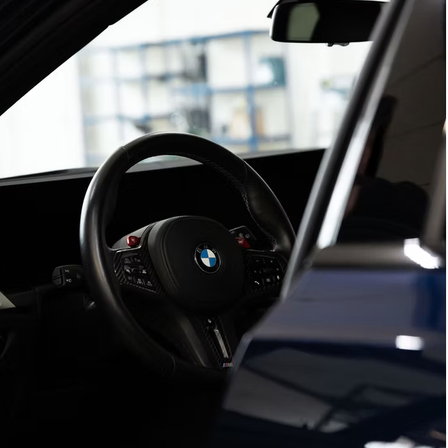 Der Innenraum eines BMW-Autos zeigt ein schwarzes Lenkrad mit roten Akzenten und einem BMW-Logo. Der Innenraum ist dunkel mit einem Glasfenster im Hintergrund.