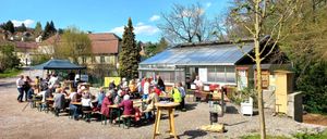 Eine Gruppe von Menschen sitzt an einem Picknicktisch vor einem Gewächshaus. Im Hintergrund befinden sich Bäume, ein Haus und ein klarer Himmel.