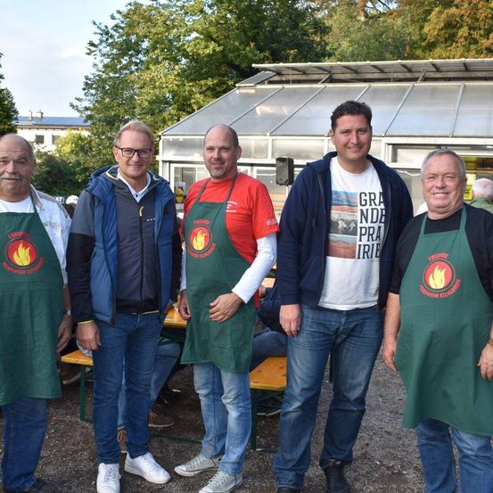 Fünf Männer in grünen Schürzen mit roten Logos stehen vor einem Gebäude. Dahinter steht ein Mann mit Brille und Jacke. Im Hintergrund befinden sich Bänke und Bäume.