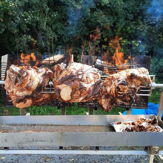 Drei große gebratene Schweine sind auf einem Metallgrill mit Flammen darunter. Das Fleisch ist gegart und hat eine knusprige Haut. Ein Tablett mit Fleischresten ist in der Nähe.