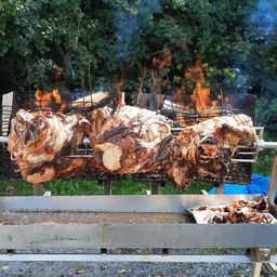 Drei große gebratene Schweine sind auf einem Metallgrill mit Flammen darunter. Das Fleisch ist gegart und hat eine knusprige Haut. Ein Tablett mit Fleischresten ist in der Nähe.