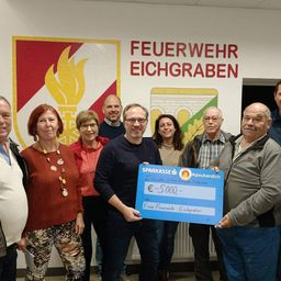 Eine Gruppe von Menschen steht vor einer Wand und hält einen blauen Scheck. Der Scheck zeigt 'Sparkasse' und '5000 Euro'. Die Leute lächeln und scheinen für ein Foto zu posieren. Hinter ihnen hat eine Wand die Worte 'Feuerwehr Eichgraben' und ein Logo.
