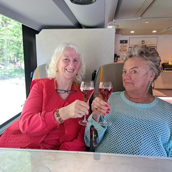 Zwei Frauen in einem Bus, eine in einem roten Top und die andere in einem blauen Top, lächeln und stoßen mit Gläsern Wein an.