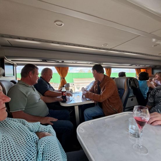 In einem Fahrzeug sitzen mehrere Personen um einen Tisch herum, unterhalten sich und trinken. Ein Mann in einer braunen Jacke hält eine Flasche. Eine Frau rechts isst und ein Weinglas steht auf dem Tisch.