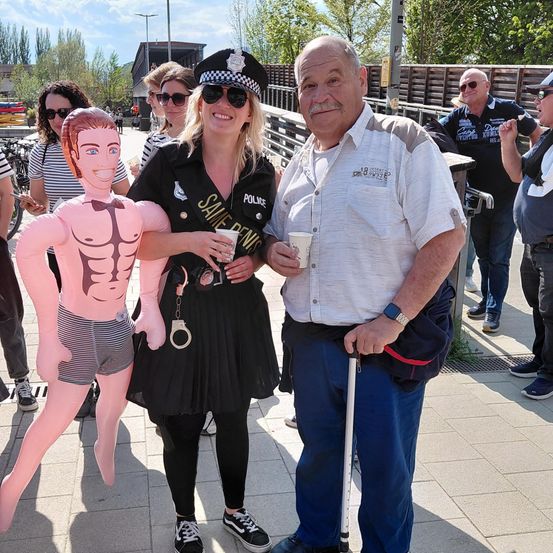Eine Gruppe von Menschen steht im Freien; ein Mann und eine Frau, die als Polizist verkleidet sind, posieren mit einer Puppe, während andere zusehen.