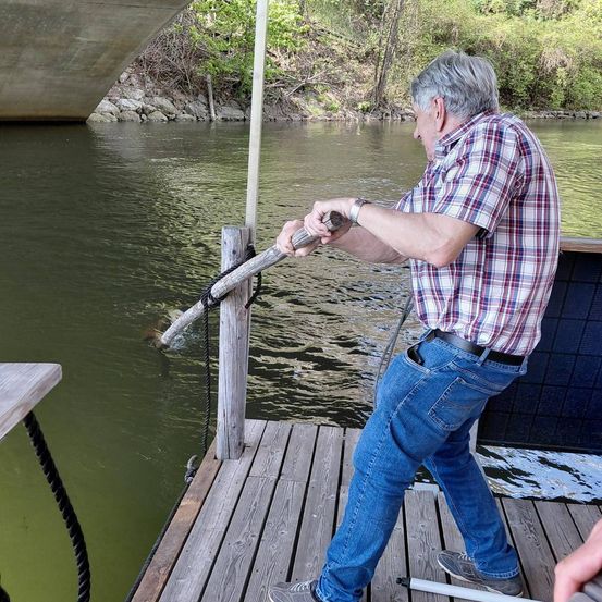 Ein älterer Mann mit einem karierten Hemd und Jeans zieht eine Seil von einem Holzsteg in der Nähe eines Flusses.