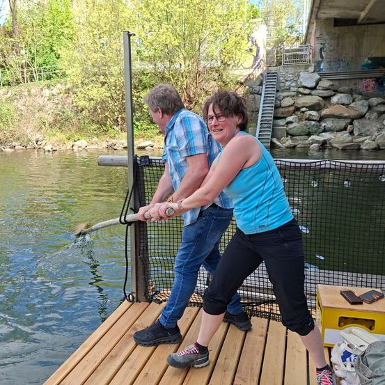 Ein Mann und eine Frau stehen auf einem Holzsteg am Wasser und halten einen langen Stock, möglicherweise zum Angeln. Hinter ihnen befinden sich Felsen, eine Treppe und eine Brücke.