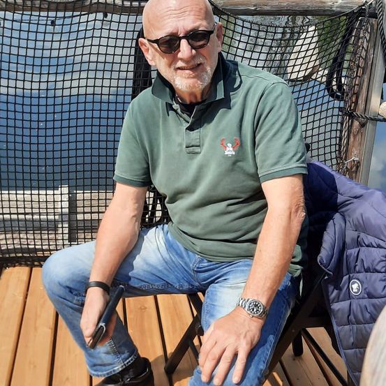 Ein älterer Mann mit Sonnenbrille und grünem Polohemd sitzt auf einer Holzterrasse mit Blick auf das Meer.