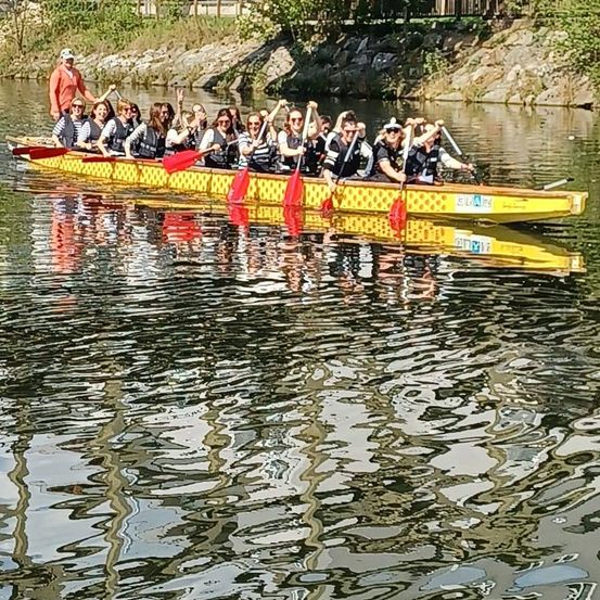 Eine Gruppe von Menschen in Schwimmwesten rudert mit einem Drachenboot auf einem Fluss, wobei eine Person in einem orangefarbenen Shirt vorne steht.