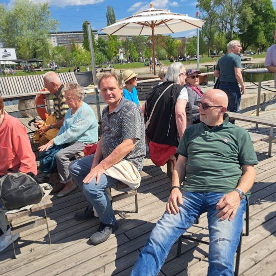 Mehrere ältere Menschen sitzen auf einer Holzterrasse in der Nähe eines Sees, einige mit Hüten und Sonnenbrillen. Ein Mann in Grün sieht entspannt aus, während andere plaudern. Ein Rettungsring und ein Regenschirm sind in der Nähe.