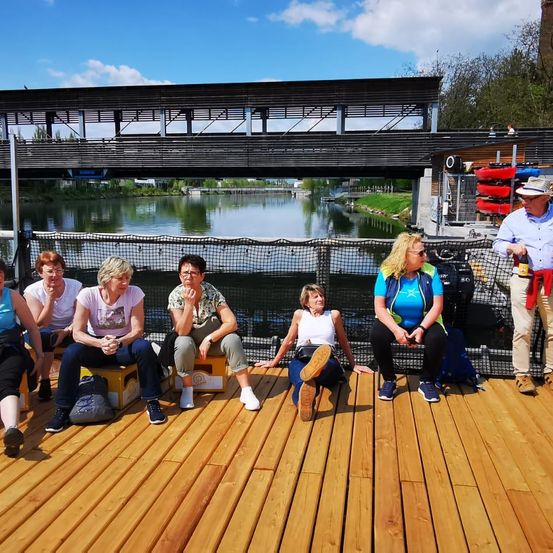 Eine Gruppe von Menschen sitzt auf einer Holzterrasse am Fluss, mit einer Brücke im Hintergrund. Eine Person hält eine Flasche, und andere haben Taschen neben sich.