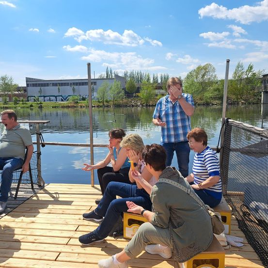 Eine Gruppe von Menschen sitzt auf einer Holzplattform über einem See, einer steht und lacht. Eine andere Frau bedeckt ihren Mund mit der Hand.
