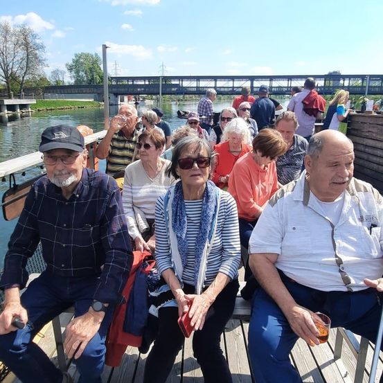 Eine Gruppe älterer Erwachsener sitzt an einer Anlegestelle am Fluss, einer trinkt. Im Hintergrund gehen Menschen über eine Brücke.