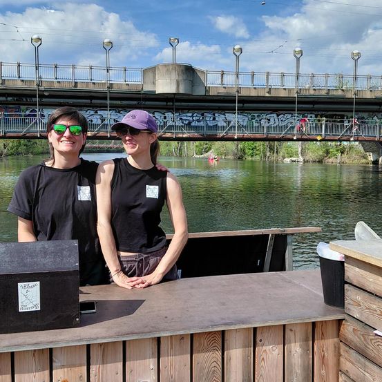 Zwei Frauen stehen an einem Holztresen am Fluss mit einer Brücke im Hintergrund. Sie tragen beide schwarze Shirts und Sonnenbrillen.