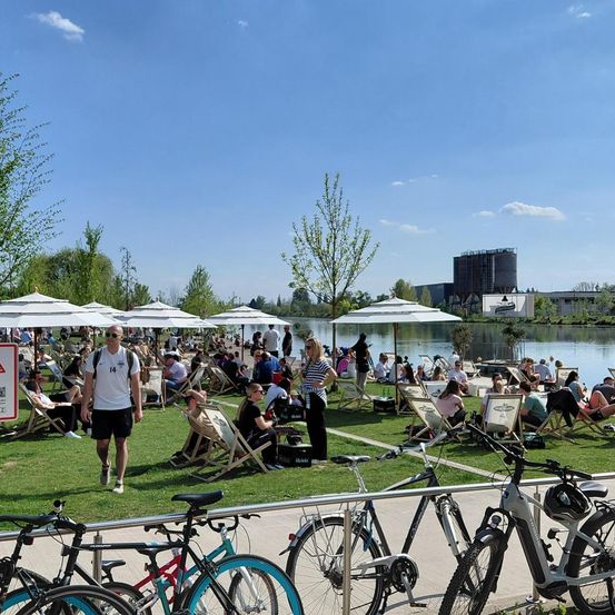 Ein sonniger Tag an einem See mit Menschen, die sich auf Stühlen unter Sonnenschirmen entspannen, einem Mann, der geht, und mehreren Fahrrädern, die an der Seite geparkt sind.