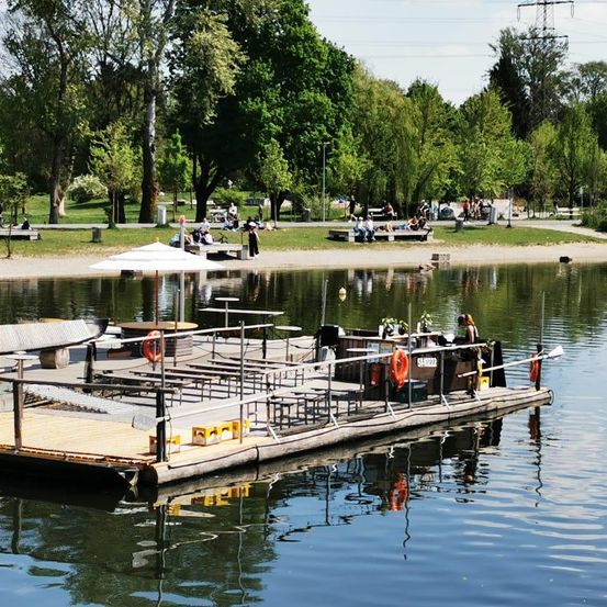 Ein Café am See mit einem Schirm und Rettungsringen auf einer schwimmenden Plattform. Leute sitzen auf Bänken und gehen am See entlang. Bäume säumen das Ufer.