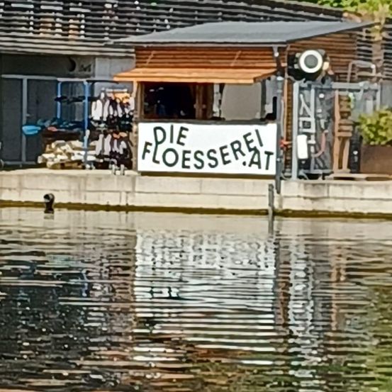 Ein kleines Holzhäuschen mit einem Vordach steht auf einem Steg. Ein Schild steht 'Die Floesserei'. In der Nähe sind ein Topf und ein Haufen Paddel zu sehen. Das Wasser reflektiert die Struktur und Umgebung.