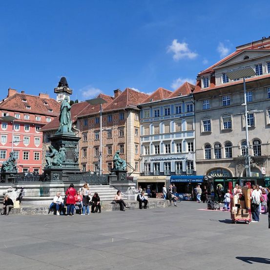 Eine Gruppe von Menschen sitzt um einen Brunnen auf einem Stadtplatz. Der Platz hat Gebäude auf allen Seiten und einen blauen Himmel.