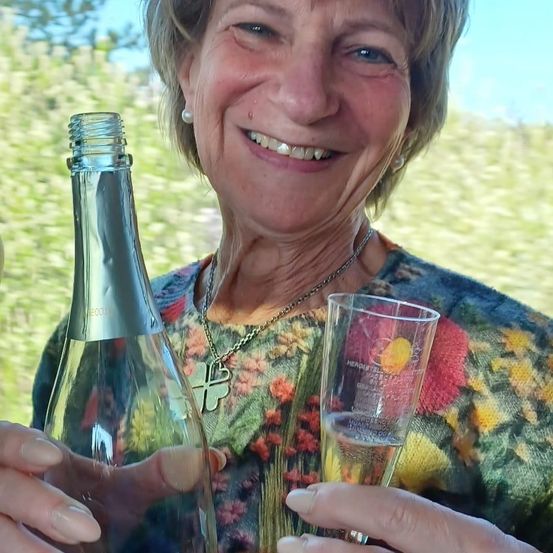 Eine ältere Frau hält eine Flasche Sekt und ein Glas und lächelt fröhlich in einer Außenumgebung.