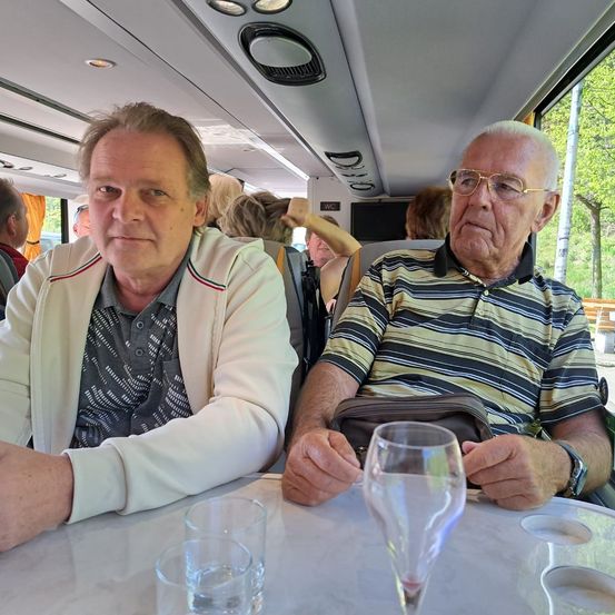 Zwei ältere Männer sitzen an einem Tisch in einem Bus, einer mit einem Weinglas. Bäume sind außerhalb des Fensters sichtbar.