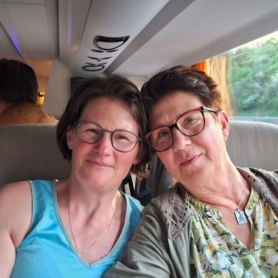 Zwei Frauen sitzen zusammen in einem Fahrzeug und lächeln für ein Foto. Eine trägt eine Brille und ein blaues T-Shirt, die andere eine Brille und ein grünes Blumenhemd. Eine dritte Person sitzt hinter ihnen.