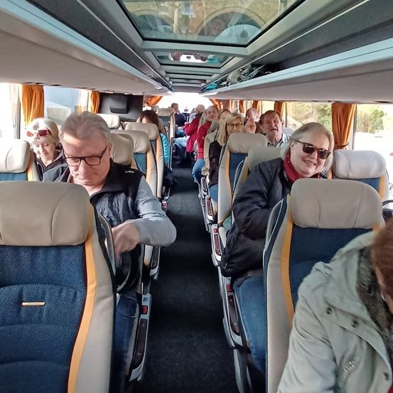 Im Inneren eines Busses sitzen Passagiere. Ein Mann mit Brille schaut nach vorne. Eine Frau vorne trägt eine Jacke und Sonnenbrille. Der Bus hat eine weiße Decke und blaue Sitze.